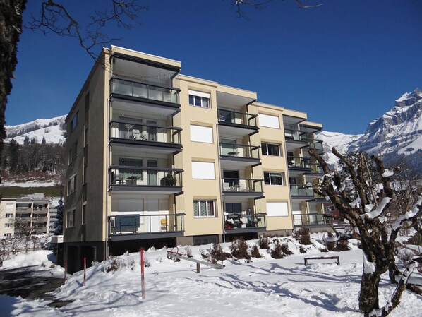 Un immeuble à plusieurs étages doté de balcons, un sol recouvert de neige et des montagnes en arrière-plan.