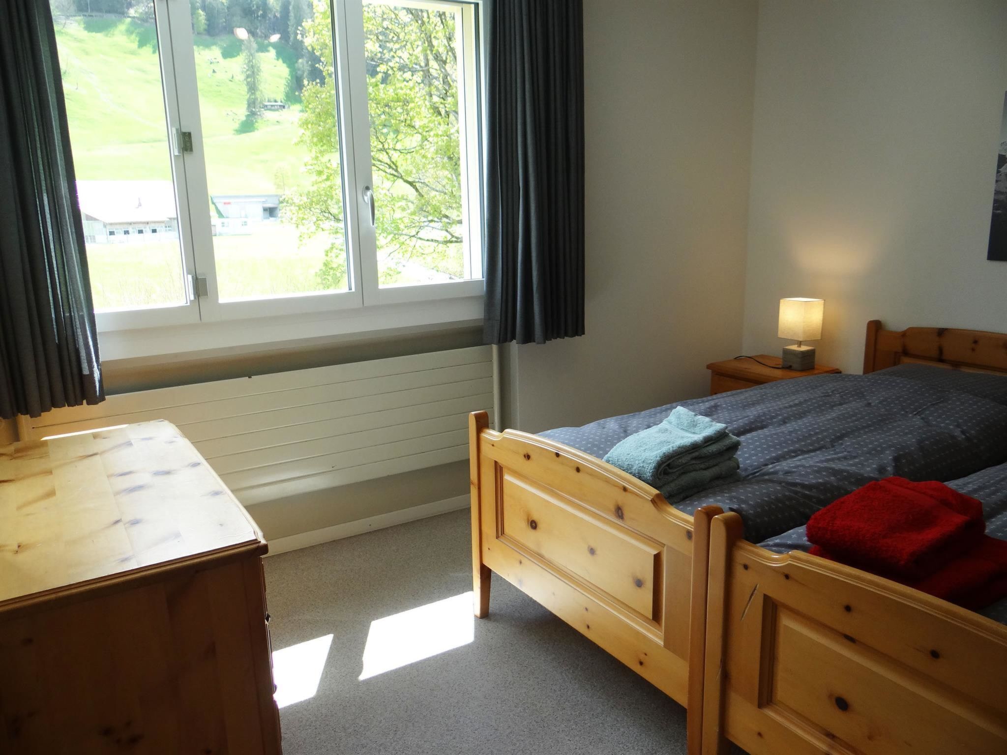 Ein Schlafzimmer mit einem hölzernen Bett, einem Nachttisch mit Lampe, einem Fenster mit Vorhängen und Blick auf eine grüne Landschaft.