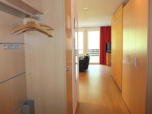 Een hotelgangen met houten vloer, een kapstok, een televisie op een kast en een balkon met rode gordijnen.