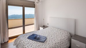 Quarto com cama, mesa de cabeceira, luminária e vista para montanhas por uma porta de correr.