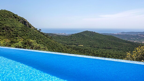 Piscina azul límpida com vista para uma paisagem montanhosa e o mar ao fundo.