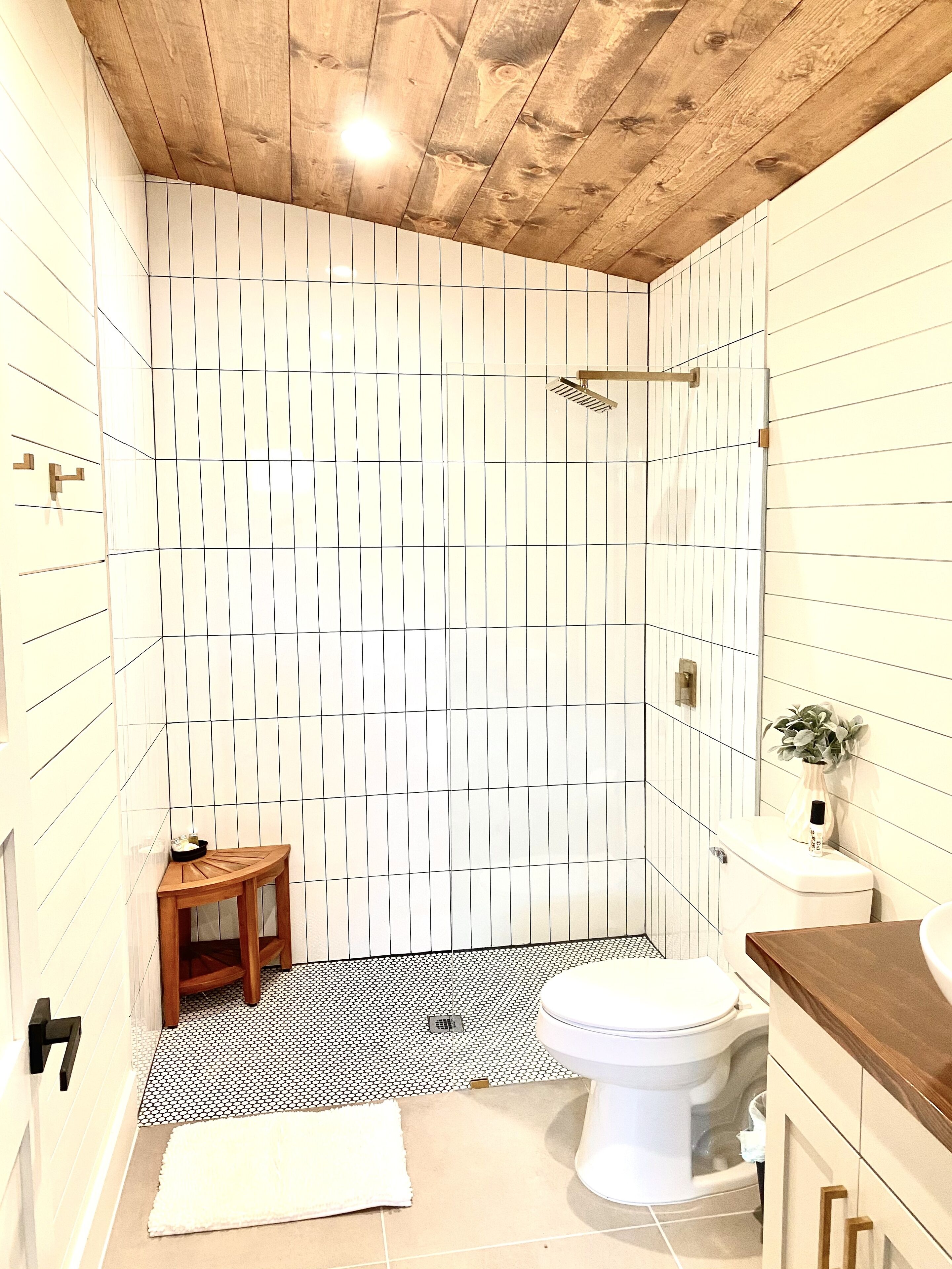 Un bagno con soffitto in legno, una parete rivestita di piastrelle bianche con motivo geometrico, pavimento a scacchi bianco e nero, un water, uno sgabello di legno e un lavandino con una pianta sul ripiano.