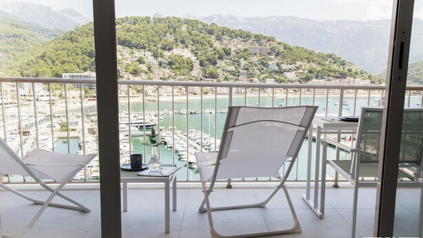 Un balcón con dos sillas blancas, una mesa y vistas a un puerto deportivo con barcos y un pueblo en la ladera.