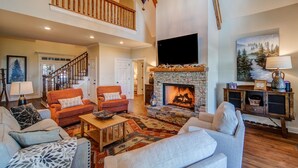 A spacious living room with a stone fireplace, a large TV, and a staircase leading to an upper level. - Big Canoe Home with Mountain Views, Game Room, Fire Pit, Sleeps 14 (Big Canoe)