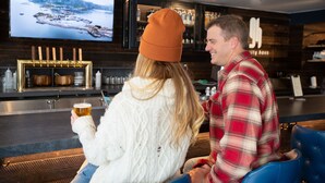 Zwei Personen sitzen in einer Barbereich mit Fernseher, der eine schöne Landschaft zeigt, einer Bar mit Flaschen und einer gemütlichen Sitzgruppe.