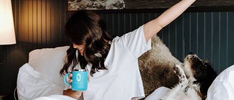 Une personne est assise sur un lit, un chien à ses côtés. Elle tient une tasse dans une main. La pièce est décorée d'une œuvre d'art murale sur le thème de la montagne et comprend une lampe.