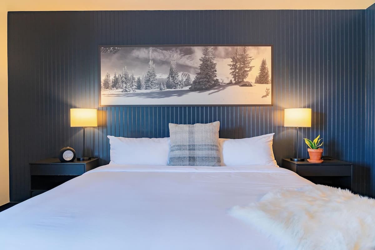 Une chambre d’hôtel avec un lit, deux lampes de chevet, un tableau accroché au mur et une plante en pot.