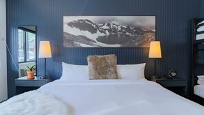 Ein Hotelzimmer mit einem großen Bett, zwei Nachttischlampen, einem Gemälde einer Berglandschaft, einer flauschigen weißen Decke und einem Nachttisch mit einer Topfpflanze.
