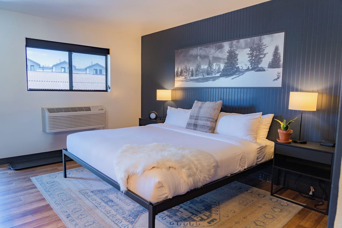 Une chambre moderne avec un grand lit, deux lampes de chevet, un climatiseur fixé au mur et une fenêtre donnant sur des immeubles.