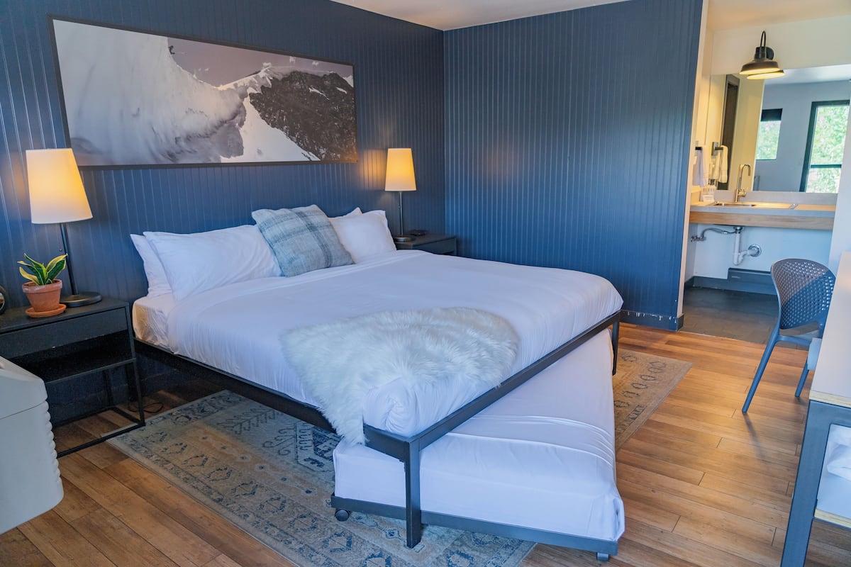 Une chambre moderne avec un grand lit, deux tables de chevet avec des lampes, une grande fenêtre et une salle de bain visible au fond.