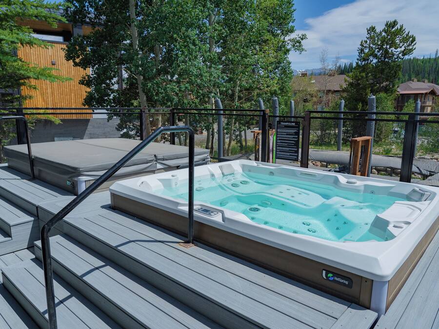 A hot tub on a wooden deck with steps leading up to it, surrounded by a metal fence and trees.