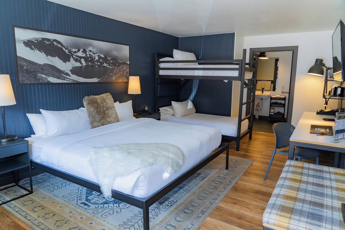 Une chambre d’hôtel avec un grand lit, un lit superposé, un bureau, une chaise et une salle de bain visible à travers une porte ouverte.