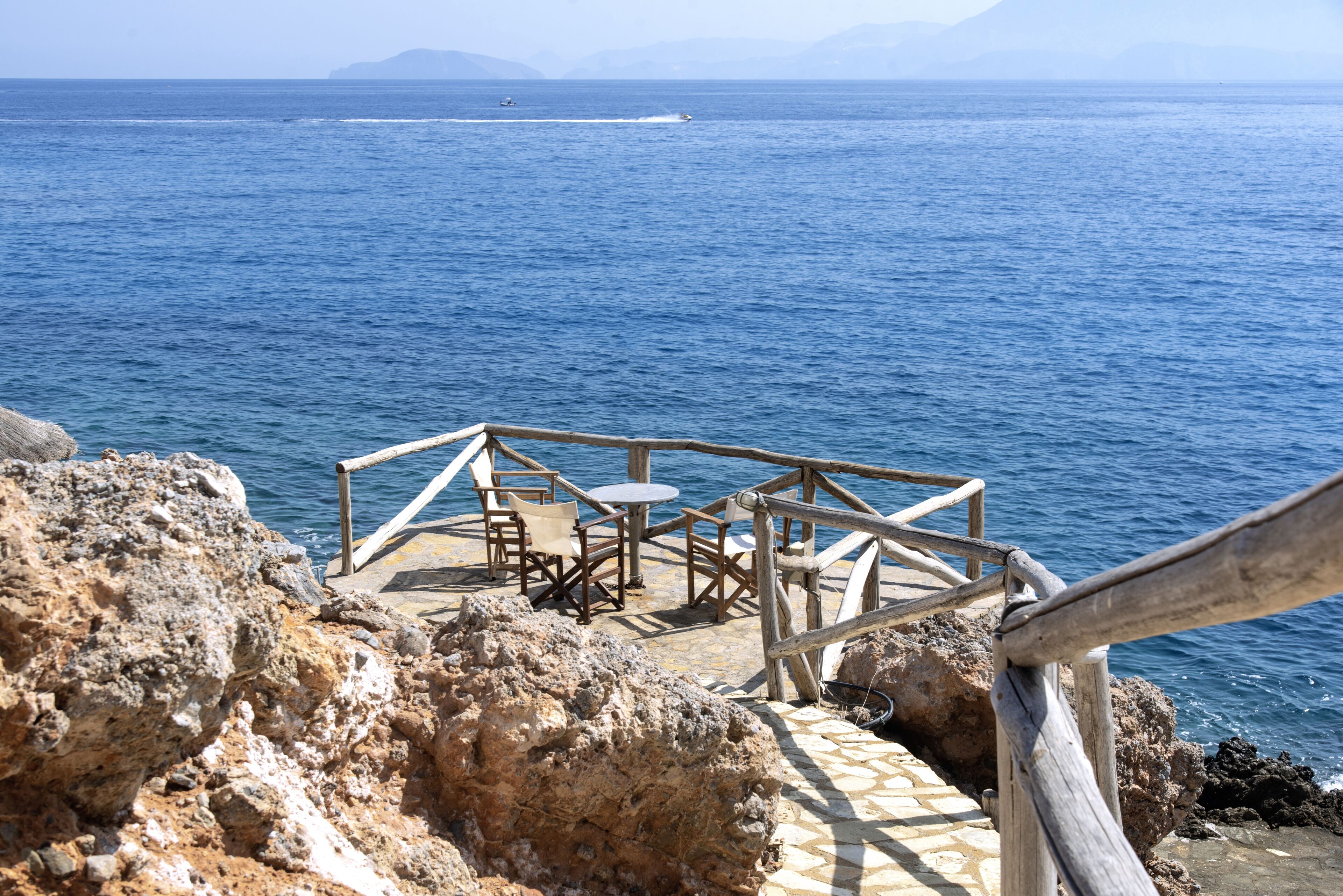 A wooden deck with chairs and tables overlooking a body of water, with rocky terrain in the foreground.