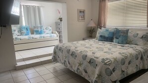 Un dormitorio con cama, televisor, cómoda, mesita de noche, lámpara y ventana con cortinas.
