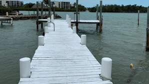 Un muelle de madera se adentra en aguas tranquilas, con edificios y vegetación al fondo.