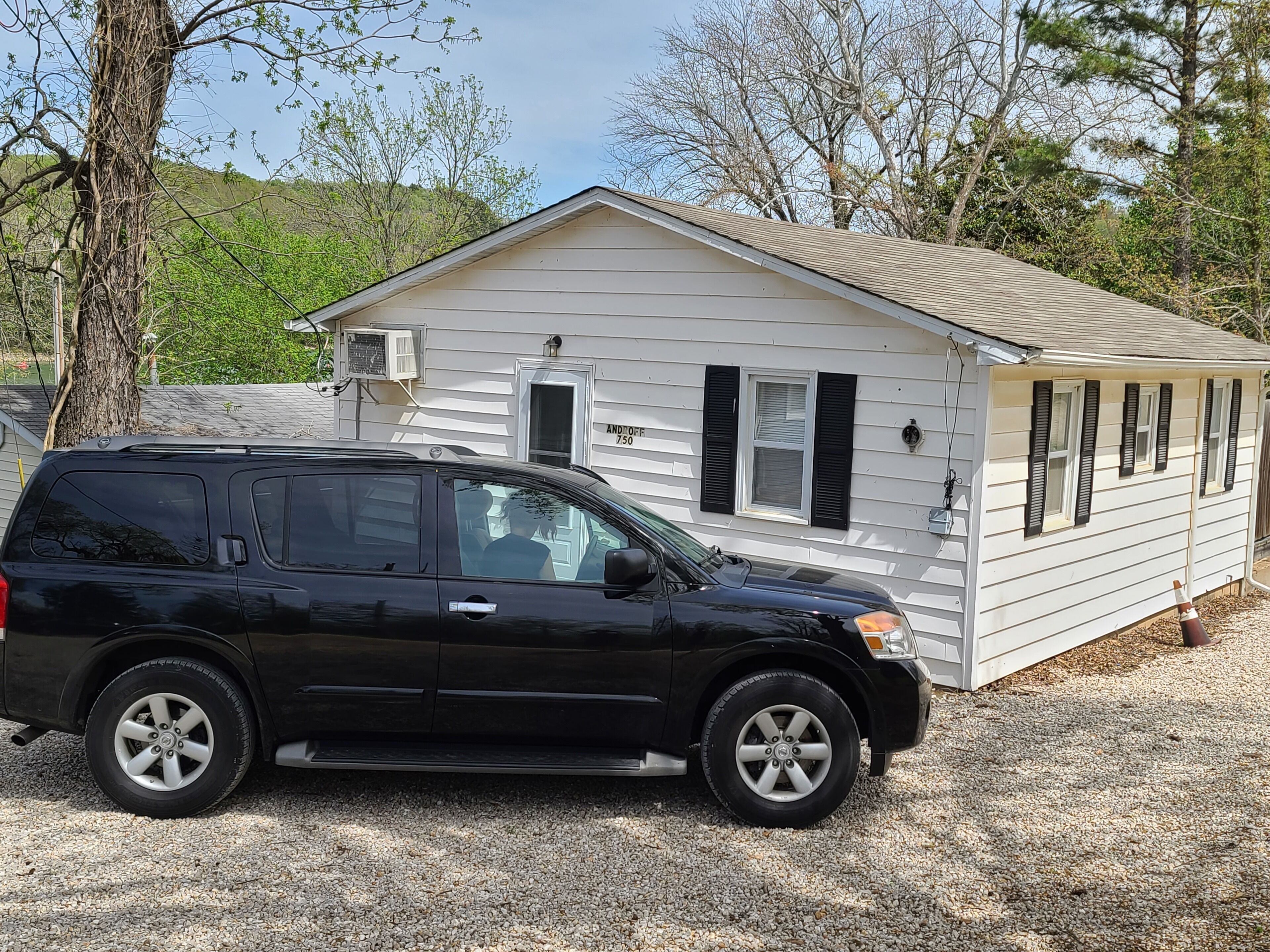 A black SUV parked in front of a white mobile home with black shutters, a gravel driveway, and trees in the background.