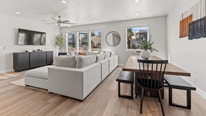 A modern living room with a grey sectional sofa, a wooden dining table with chairs, a ceiling fan, and a large window offering a view of the outside.