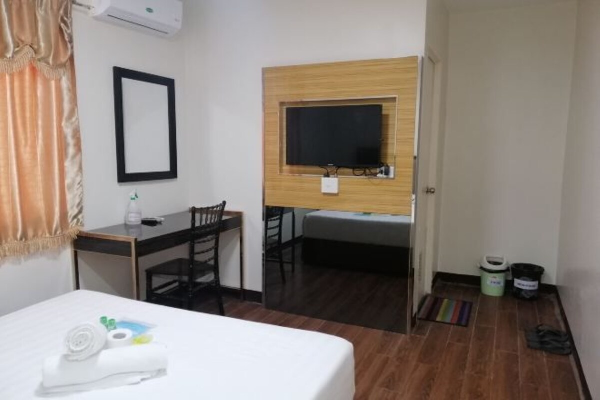 Habitación de hotel con una cama, un escritorio, una silla, un televisor fijado a un panel de madera, una puerta y un cubo de basura pequeño.