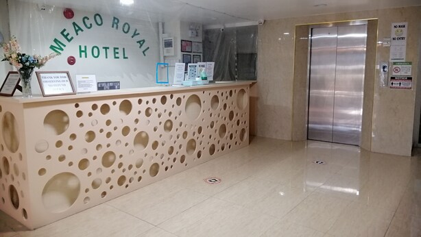 Una mesa de recepción con un letrero que dice 'Meaco Royal Hotel', un aviso de política sin mascarilla y un letrero de prohibido el acceso.