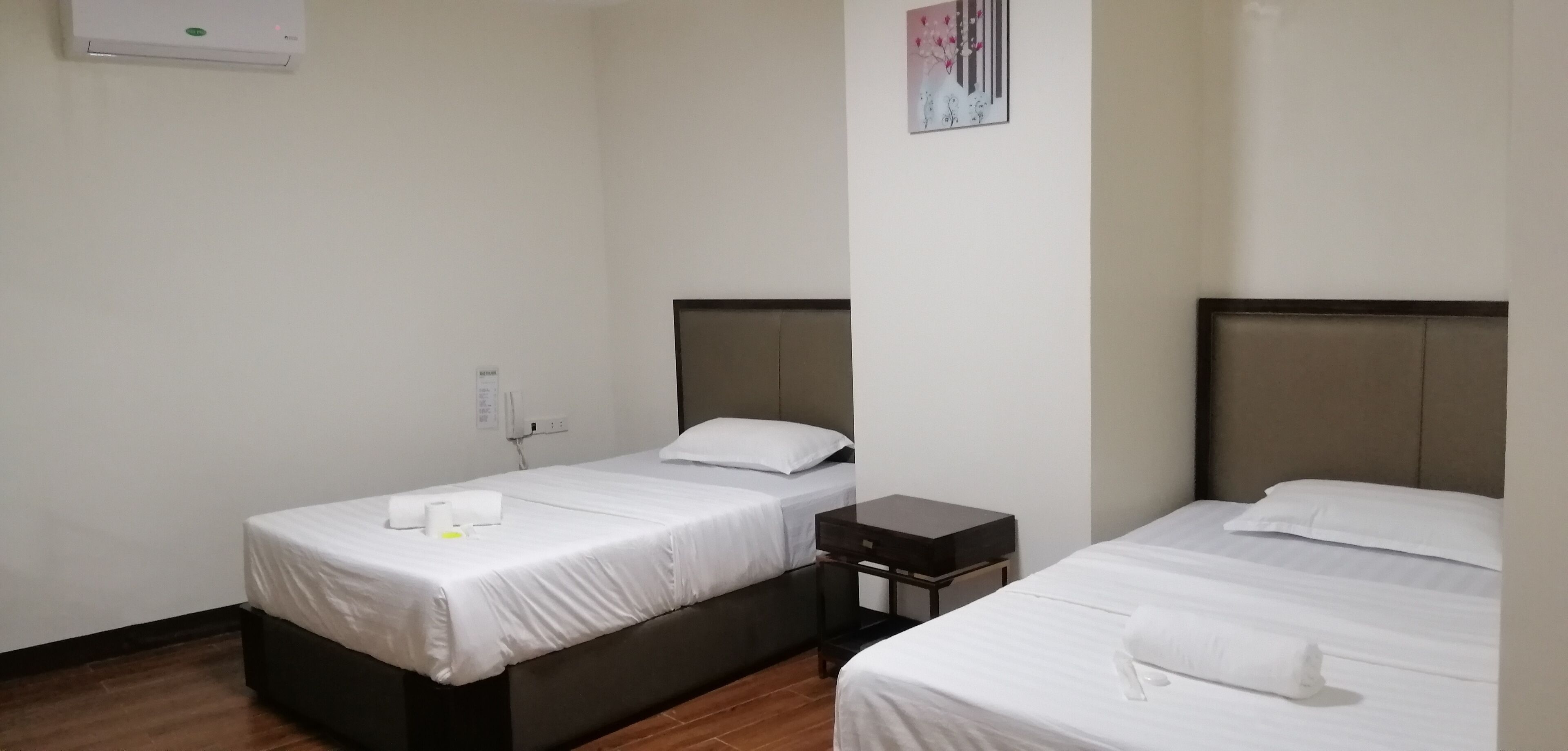A hotel room with two single beds, a small bedside table, a wall-mounted air conditioner, and a framed picture on the wall.