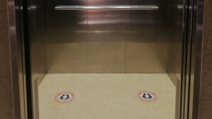 Un ascensor con interior metálico, un letrero de seguridad y dos indicaciones de salida de emergencia en el suelo.