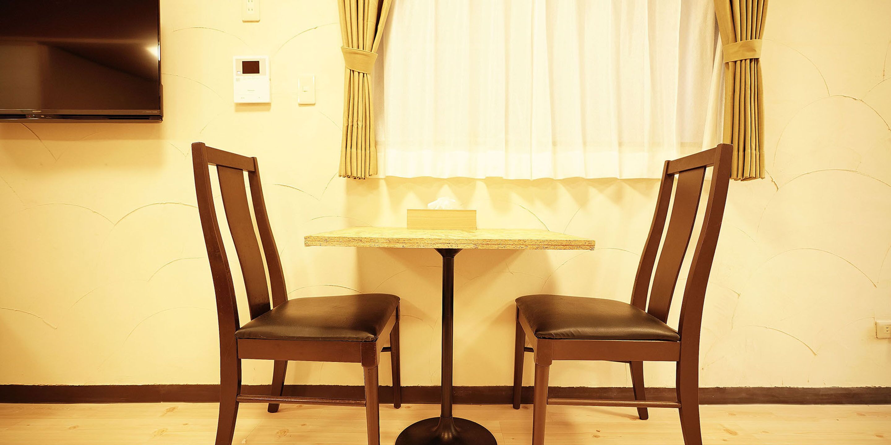 A room with two wooden chairs, a small round table, a flat-screen TV mounted on the wall, and a window with curtains.