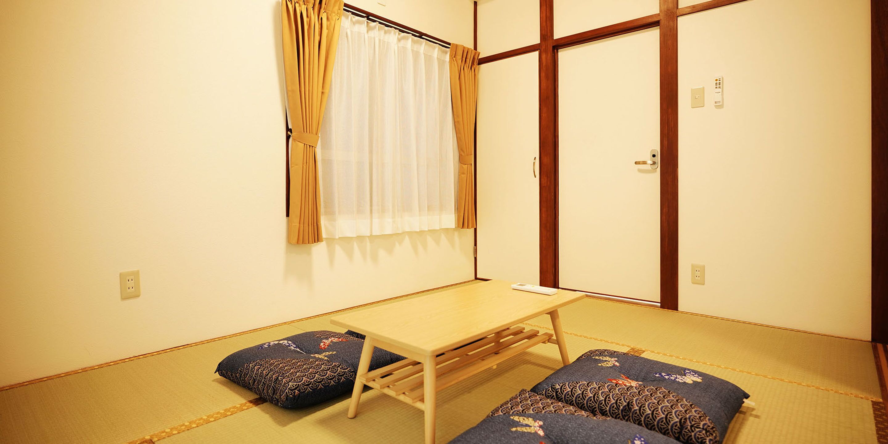 A traditional Japanese room with tatami flooring, a low wooden table, two tatami mats with cushions, a sliding door, and a window with curtains.