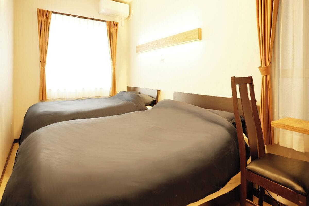 A hotel room with two beds, a wooden chair, a nightstand, and a window with curtains.