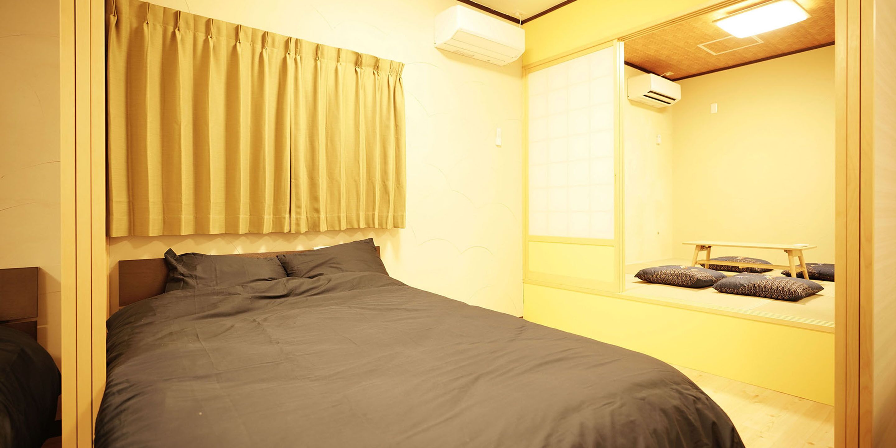 A bedroom with a bed, curtains, an air conditioning unit, a mirror, and a bench with pillows.