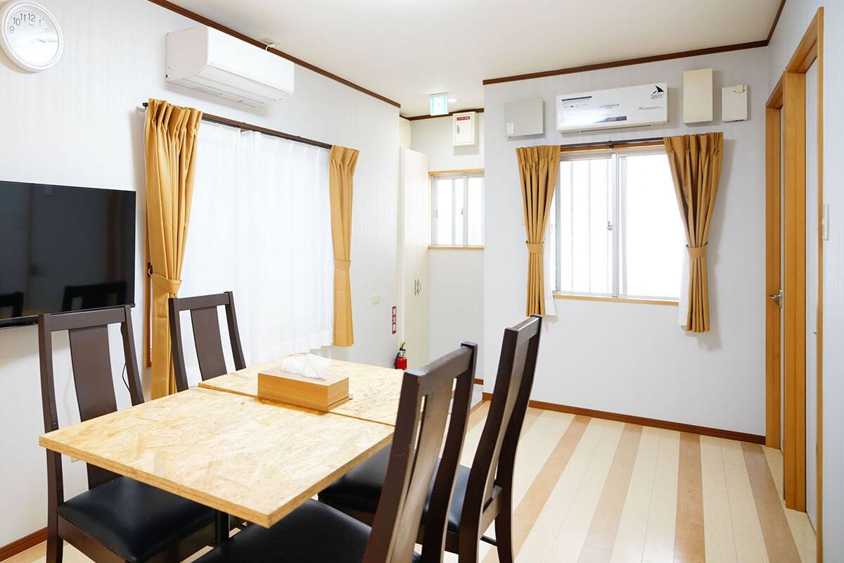 A dining area with a wooden table and chairs, a flat-screen TV mounted on the wall, and two windows with curtains.