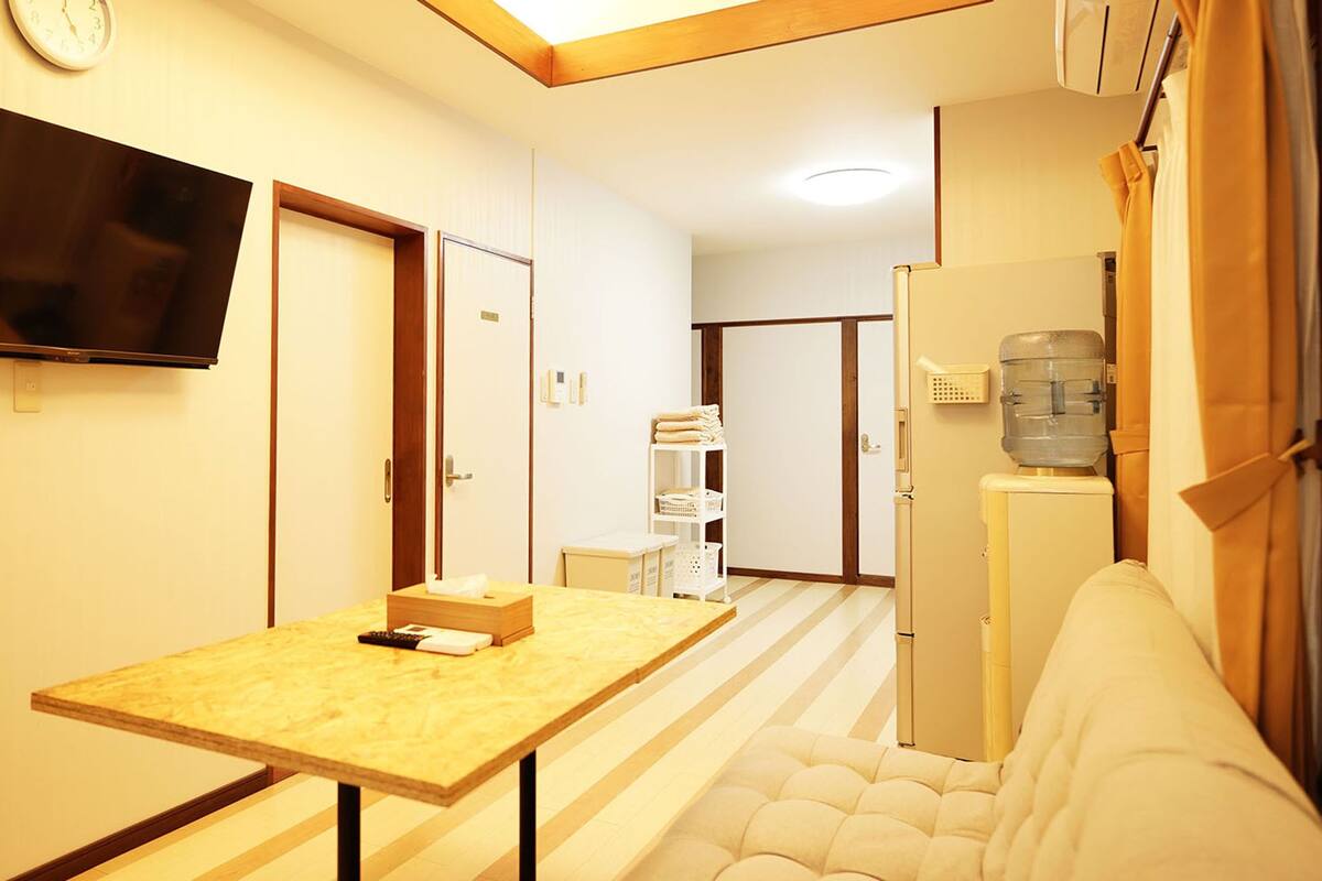 A hotel room with a wooden table, a sofa, a TV mounted on the wall, and a water dispenser.