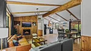 A spacious living area with a fireplace, wooden beams, a dining table, and a kitchen in the background. - Creekside Cabin~Jacuzzi~fire pit~screened porch (Cosby)