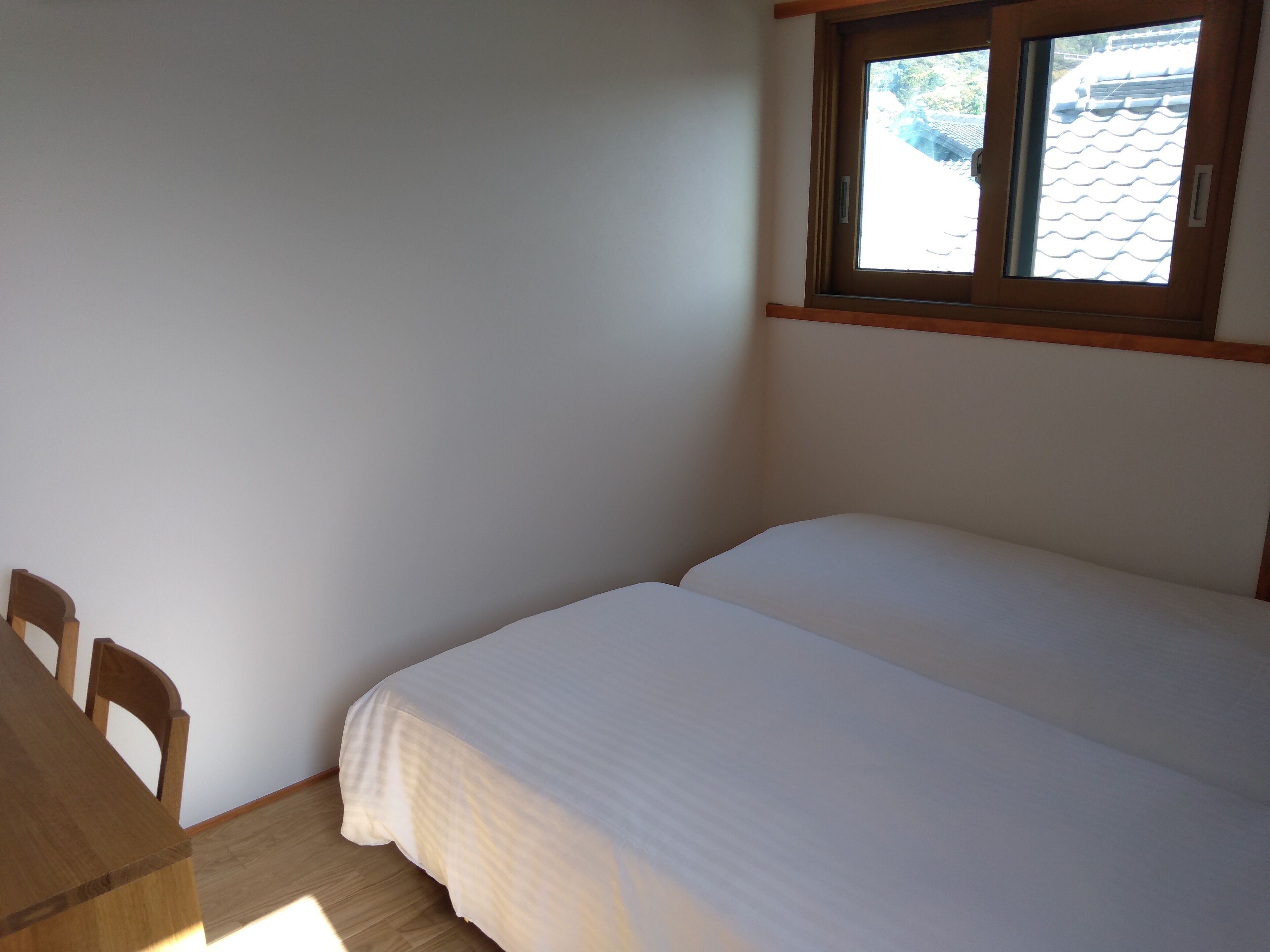 A small, clean room with a single bed, a wooden table, and a window with a view of a tiled roof.