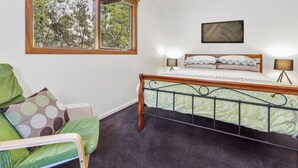 A bedroom with a bed, two bedside lamps, a window with a view of trees, a chair with a cushion, and a framed picture on the wall. - Blackwood Chalet - Base of Buller (Sawmill Settlement)