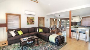 A modern living room with a sectional sofa, a coffee table, and a patterned rug. There is a dining area with a wooden table and chairs, and a kitchen with a bar area. - Blackwood Chalet - Base of Buller (Sawmill Settlement)