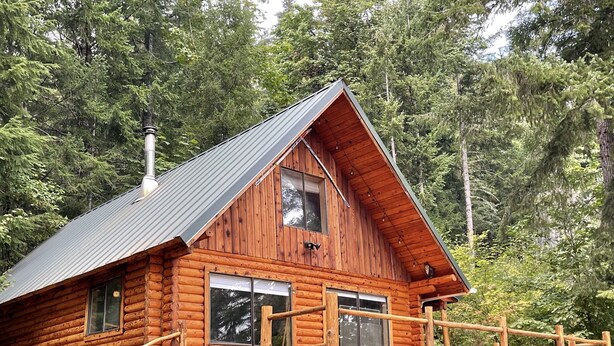 A log cabin with a metal roof, wooden deck, and a staircase leading to the entrance. - Woolly Bear Log Cabin Mountain Get-Away (Packwood)