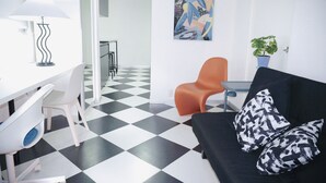 Ein modernes Zimmer mit einem schwarz-weißen Schachbrettboden, einer schwarzen Couch mit gemusterten Kissen, einem weißen Schreibtisch mit Lampe und einem blauen Blumentopf.