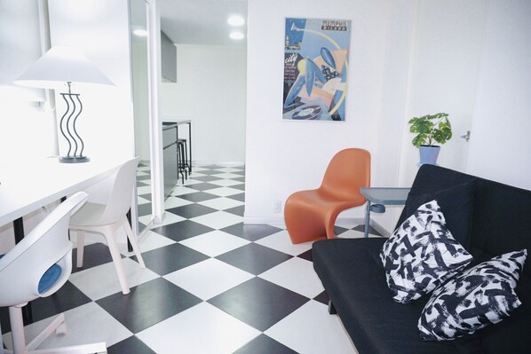 A modern room with a black and white checkered floor, a black sofa with patterned cushions, a white desk with a lamp, and a blue potted plant.