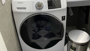 Eine kompakte, weiße Samsung Waschmaschine mit digitaler Steuerung in einem kleinen Waschraum mit grauen Fliesenwänden und einem Schachbrettfußboden.