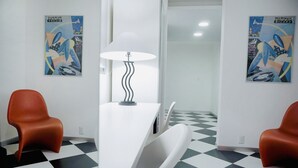 Ein Zimmer mit einem weißen Schreibtisch, zwei orangefarbenen Stühlen, einer weißen Lampe und zwei abstrakten Kunstwerken an den Wänden.