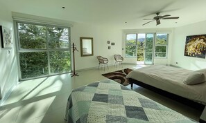 Ein Schlafzimmer mit zwei Betten, großen Fenstern, einem Deckenventilator und Blick auf Grünfläche.