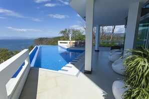 Ein Poolbereich im modernen Stil mit Meerblick, umgeben von weißen Wänden und einem Balkon mit Sitzgelegenheiten.
