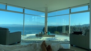 A spacious room with large floor-to-ceiling windows, a central seating area with a coffee table, and a bed with a view of the ocean and mountains.