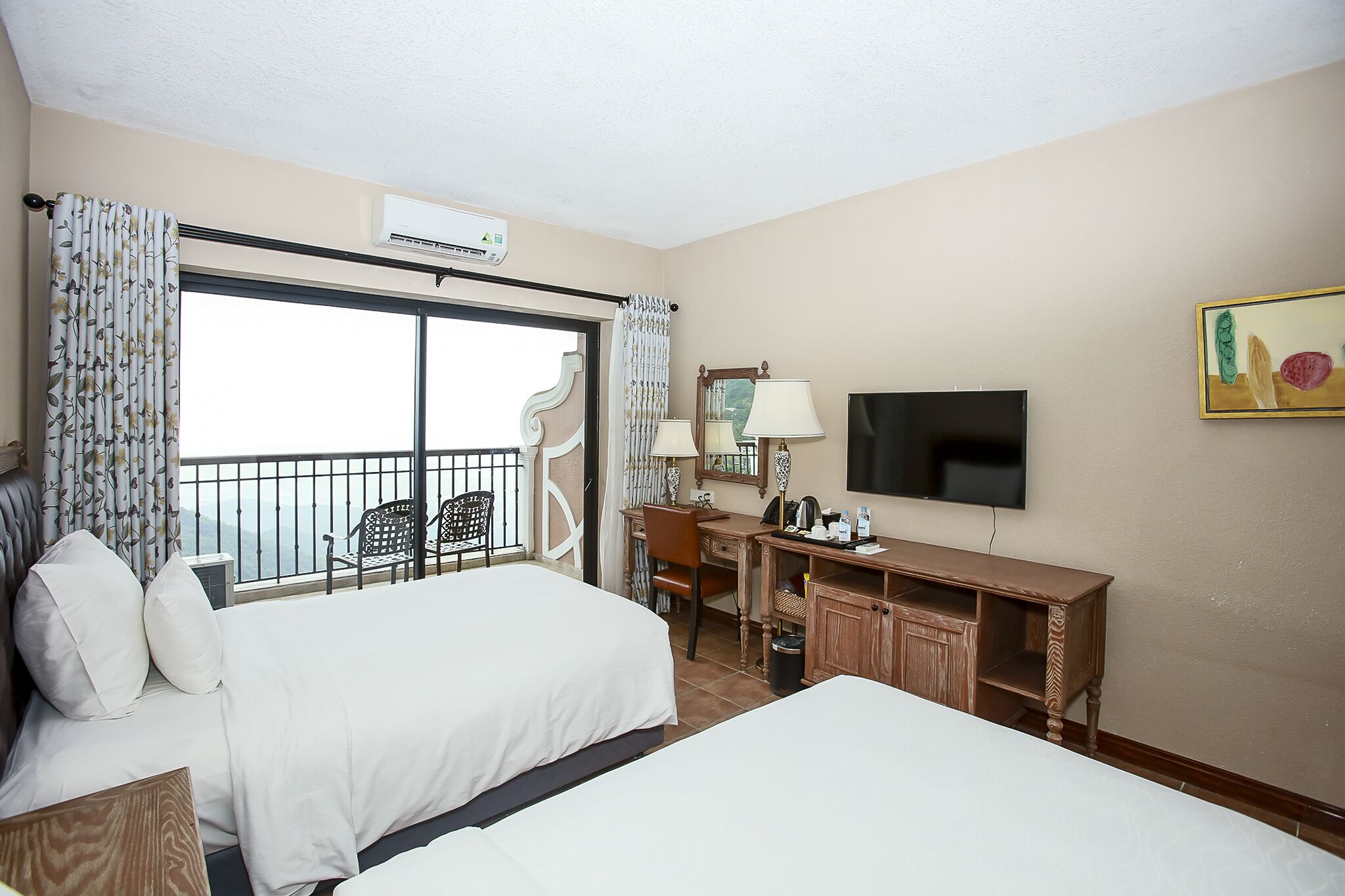 A hotel room with two beds, a desk, a chair, a TV, and a balcony with a view.