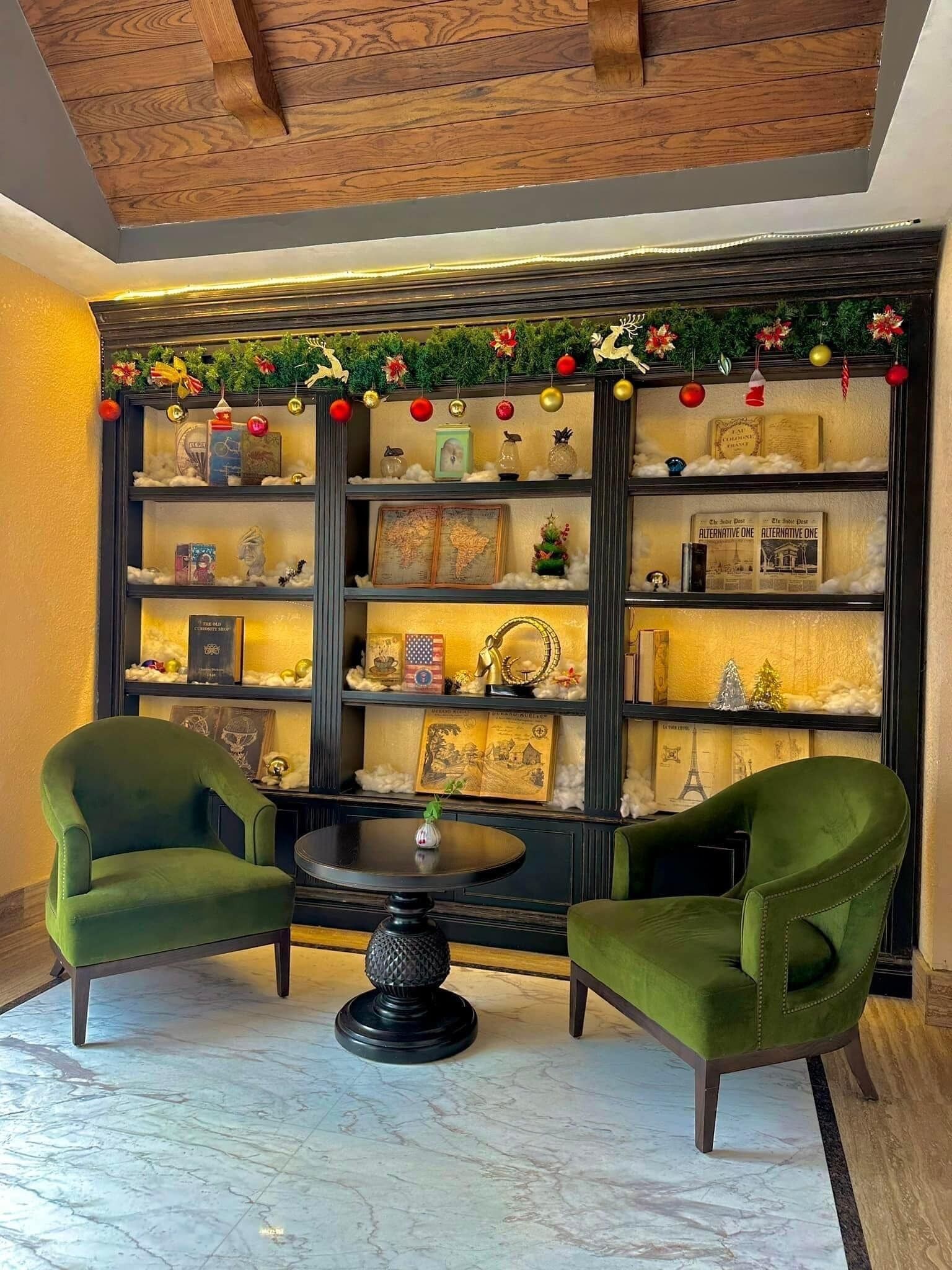 Una habitación con techo de madera, una estantería con diversos objetos decorativos, dos sillones verdes y una mesa redonda con un jarrón.