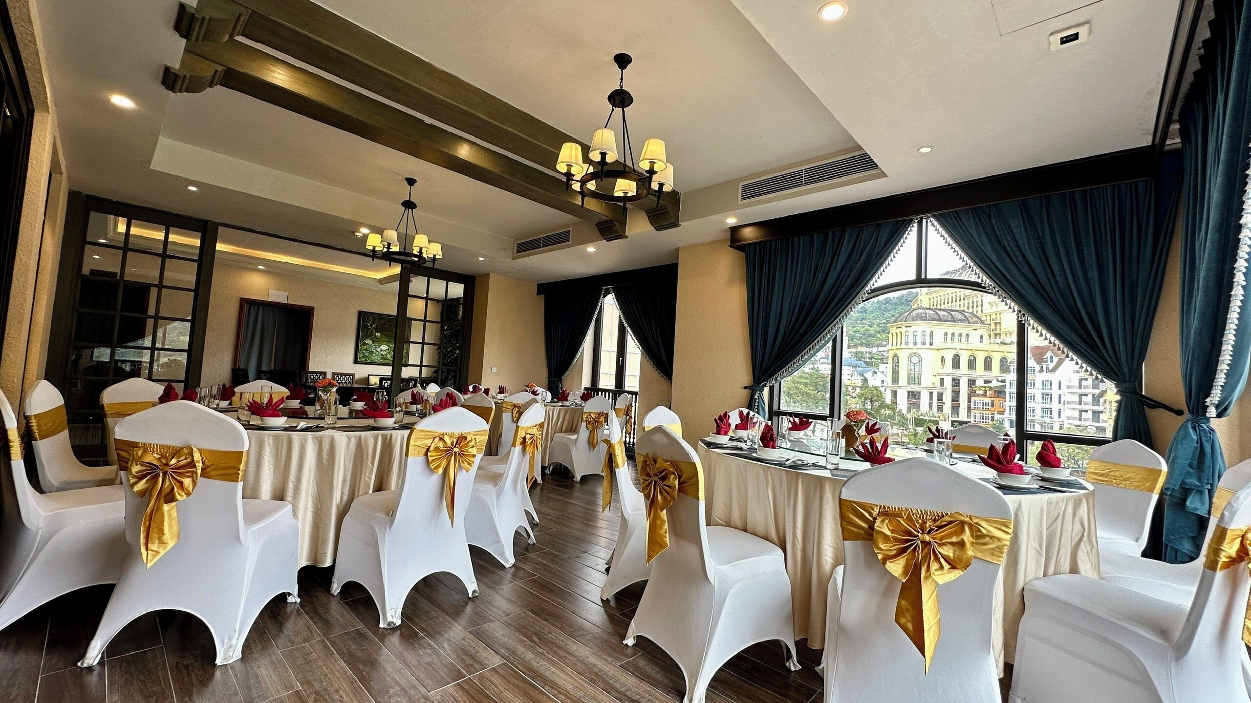 Une salle de banquet avec des tables rondes, des chaises blanches ornées de rubans dorés et un lustre.