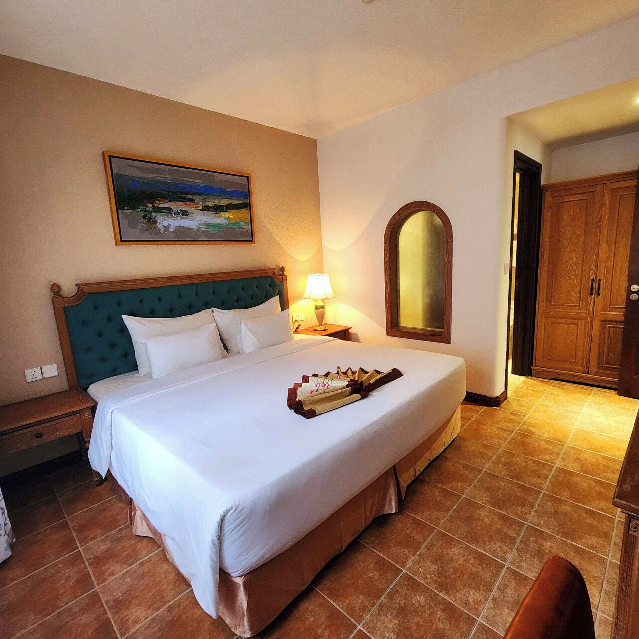 A hotel room with a large bed, a nightstand, a lamp, a painting, and a wardrobe.
