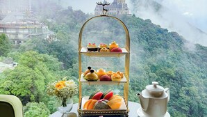 Une table sur laquelle est disposé un présentoir à étages pour les desserts, une théière, ainsi que divers gâteaux et confiseries, le tout mis en valeur par un paysage montagneux luxuriant et un château en arrière-plan.