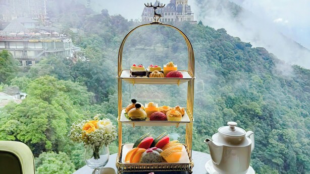 A table with a tiered dessert stand, a teapot, and various pastries and sweets, set against a backdrop of a lush mountain landscape and a castle. - Venus Hotel Tam Dao (Tam Dao)