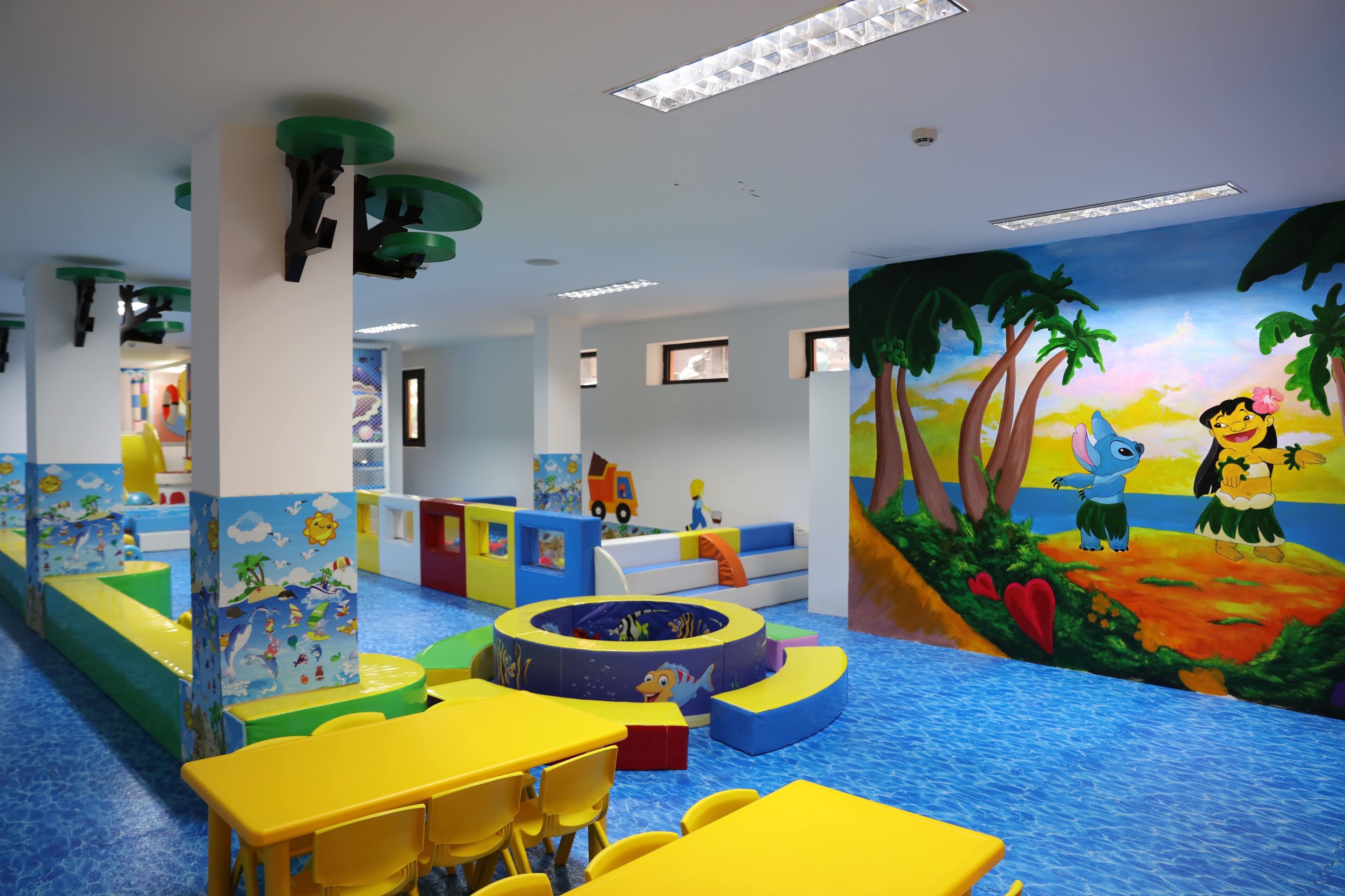 Zona de juegos infantiles con mobiliario colorido, un mural y un tapete azul.